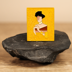 Small rectangular yellow and gold enamel pin with a reimagined gemoetric portrait of Adele Bloch-Bauer I by Gustav Klimt, with gold glitter, placed on a dark stone with a beige background.